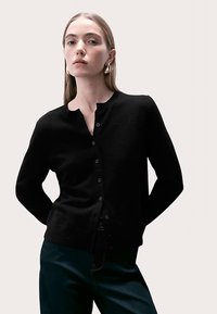 Czarny kardigan wykonany z miękkiego materiału, z okrągłym dekoltem, zapięciem na guziki z przodu oraz długimi rękawami. Model stoi na neutralnym tle.