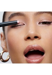 Nahaufnahme eines Gesichts, das einen rosa Lidschattenstift aufträgt. Hat glatte Haut, hellbraune Augen und glänzende nudefarbene Lippen, sowie große Creolen.