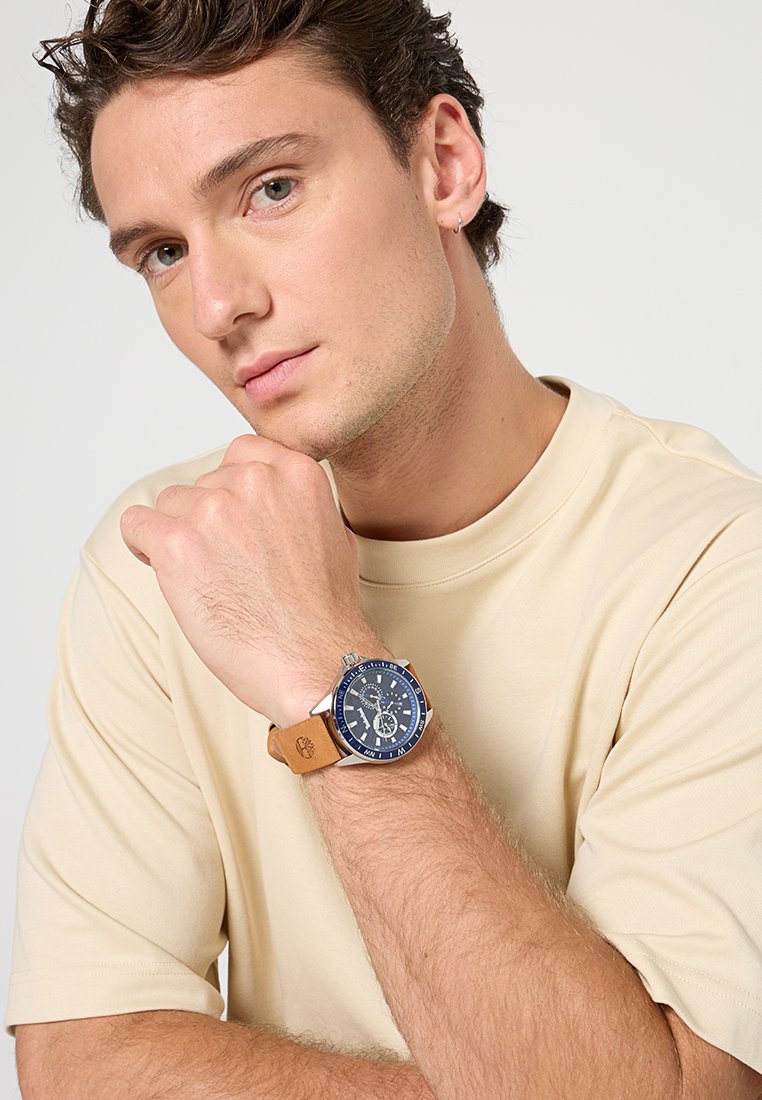 Reloj con una esfera azul marino y acentos plateados, correa de cuero marrón y una caja metálica redonda. Diseño simple y funcional.