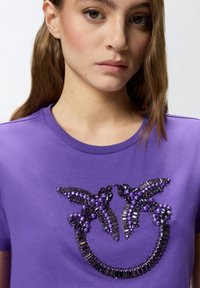 Ljubičasta pamučna majica s ukrasnim perlicama u različitim nijansama ljubičaste i crne boje oko izreza, stvarajući teksturirani naglasak.