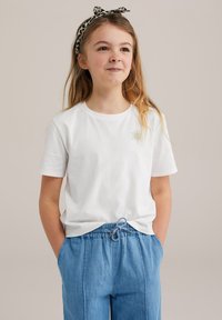 Camiseta blanca de algodón con un pequeño diseño de sol dorado en el lado izquierdo del pecho, combinada con pantalones de tiro bajo azul claro con cordón. Textura suave, ajuste casual.