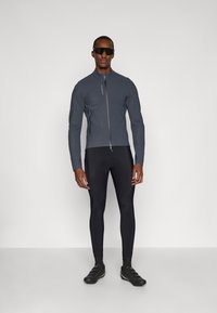 Chaqueta deportiva gris con un alto cuello, cierre de cremallera y tirador de cremallera en contraste. Combinada con mallas negras ajustadas y zapatos de ciclismo.