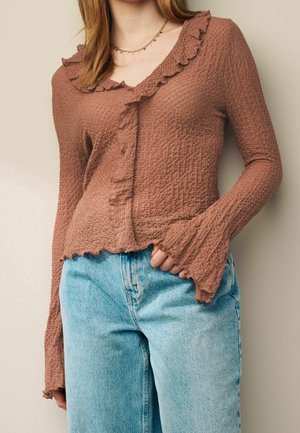 Kvinde iført en tekstureret brun langærmet top med flæsekant kombineret med lyseblå jeans og en fin guldhalskæde.