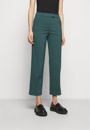 Osoba nosząca turkusowe skrócone proste spodnie, czarne mokasyny i czarny bezrękawnik na jednolitym białym tle.