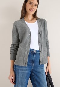 Cardigan grigio a maglia con chiusura a bottoni, polsini a costine e vestibilità rilassata, indossato sopra una t-shirt bianca e abbinato a jeans in denim blu.