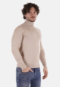 Maglione beige a collo alto in un morbido tessuto knit, design aderente, polsini e orlo a coste, indossato su jeans blu. Tessuto liscio, dettagli minimi.