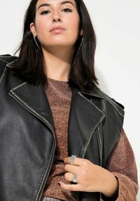 Schwarze Leder-Bikerweste über einem metallic bronzefarbenen Oberteil. Das Model trägt silberne Ringe und Kreolen. Glatte Textur, geometrische Ringdesigns.