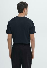 Uomo con capelli corti scuri che indossa una maglietta nera a maniche corte e pantaloni neri, visto da dietro su uno sfondo semplice.