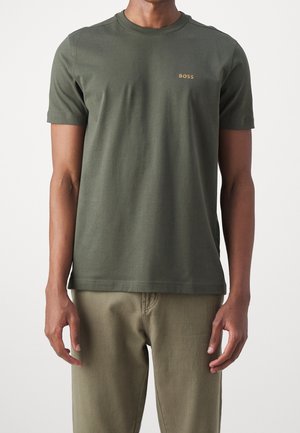 Man draagt een olijfgroene korte mouw T-shirt met een klein geel "BOSS"-logo op de linkerborst en een bijpassende olijfgroene broek.