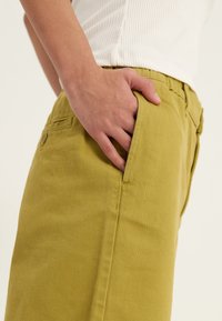 Pantalons en coton jaune à coupe droite, avec poches latérales et détails de couture visibles. Le tissu a une finition douce et texturée.
