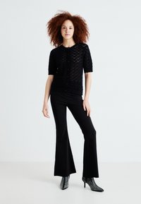 Cardigan noir en tricot à manches courtes, avec un motif texturé, associé à un pantalon noir évasé et des bottines noires à clous.
