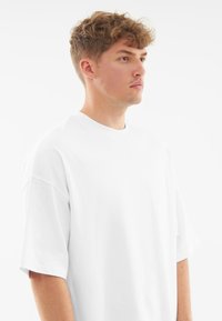Bílý oversized bavlněný tričko s kulatým výstřihem, krátkými rukávy a hladkou texturou, předváděné na modelce na jednobarevném pozadí.