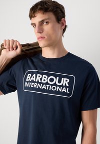 Μπλέ βαμβακερό t-shirt με μεγάλο λευκό λογότυπο "BARBOUR INTERNATIONAL". Το t-shirt έχει στρογγυλή λαιμόκοψη και κοντά μανίκια.