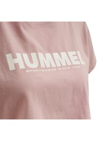 Camiseta de algodón rosa con el texto blanco "HUMMEL" y "SPORTSWEAR SINCE 1923". Cuello redondo con mangas cortas. Textura suave.