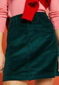 Groene corduroy minirok met verticale ribbels, voorzien van twee steekzakken aan de voorkant en een aangesloten taille, gecombineerd met een roze top met lange mouwen.