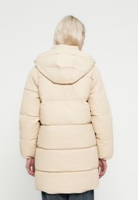 Casaco acolchoado bege com capuz, apresentando um design acolchoado e punhos elásticos. Exterior liso com um corte de comprimento médio.