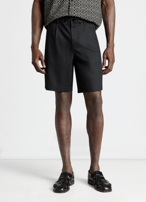 Uomo con pantaloncini neri con coulisse, scarpe nere in pelle intrecciata e top senza maniche con motivo, in piedi su uno sfondo bianco semplice.