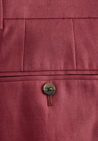 Gros plan sur un pantalon en tissu marron montrant la poche arrière avec les détails de couture et un seul bouton rond foncé.