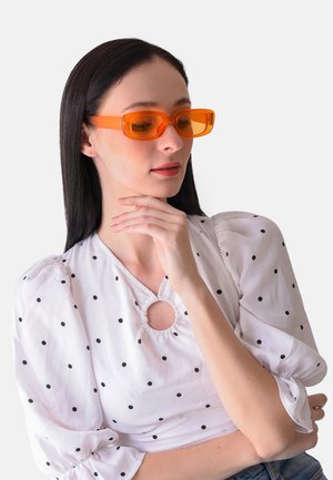 Orange rechteckige Sonnenbrille mit transparentem Rahmen. Das Modell trägt eine weiß gepunktete Bluse mit Puffärmel. Rundhalsausschnitt-Detail.