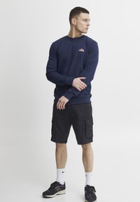 Jūrinės spalvos džemperis su mažu saulės logotipu, derinamas su juodomis krovinių šortais. Modelis užsidėjęs juodus sportbačius ir baltas kojines, stovi prieš baltą foną.