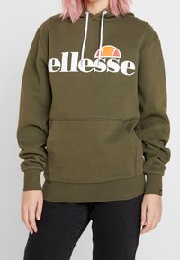 Olivgrön hoodie med en vit "ellesse" logotyp och grafiken av en orange sol, med en framficka och ribbade muddar och nederdel.