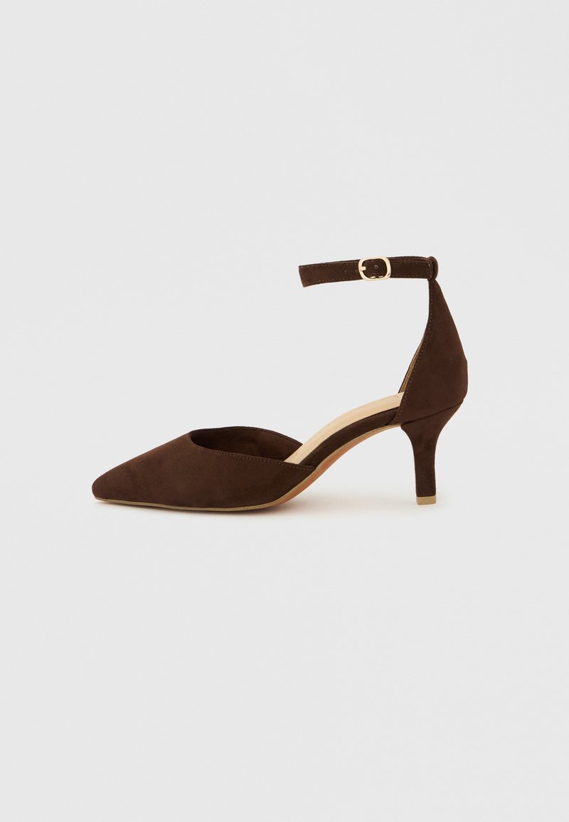 Escarpin à talon bas en daim marron avec un bout pointu et une bride de cheville munie d'une boucle ronde. Texture lisse et design minimaliste.