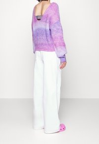 Lila ombré-tröja med V-hals och draperade axlar, tillsammans med vita vida byxor och rosa träskor, som visar en texturerad stickning.