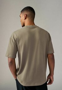 Camiseta caqui de manga corta, corte relajado, cuello redondo y textura suave, combinada con una cinturilla negra. Vista desde la parte posterior.