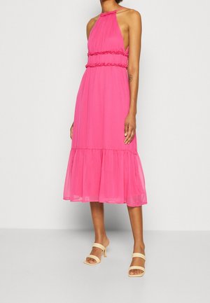 Kobieta w różowej sukience midi bez rękawów z falbankami i beżowych sandałach na obcasie, stojąca na jednolitym tle.