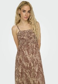 Robe maxi de couleur beige clair avec un motif de feuilles marron. Dotée de bretelles spaghetti ajustables et d'un corsage froncé pour un ajustement décontracté.