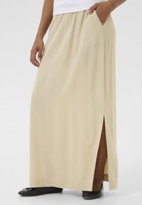 Jupe maxi beige avec fente latérale et ceinture froncée, fabriquée en tissu léger. Le modèle porte des chaussures plates noires.