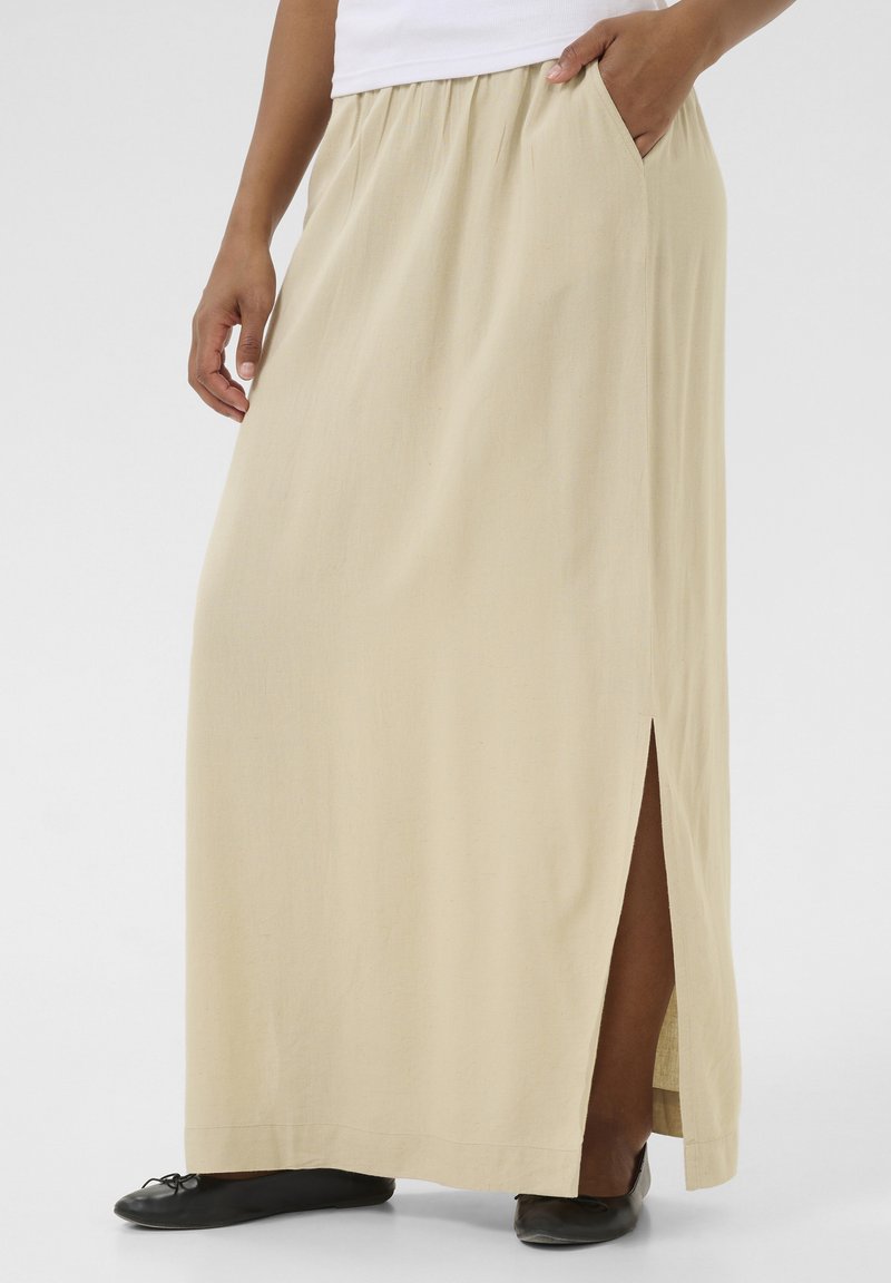 Jupe maxi beige avec fente latérale et ceinture froncée, fabriquée en tissu léger. Le modèle porte des chaussures plates noires.