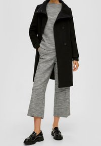 Černý kabát s vysokým límcem přes texturovaný šedý svetr a odpovídající široké kalhoty. Lesklé černé boty dokončují tento outfit.