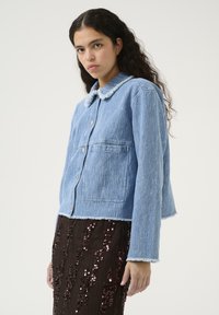 Giacca di jeans blu con colletto peloso, bottoni a pressione, due tasche frontali e orli grezzi. Indossata sopra una gonna marrone con paillettes.