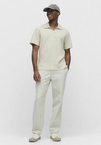 Paita polo dalam kain Beige muda dengan tekstur, dipadukan dengan celana Beige muda, topi abu-abu, dan sepatu sneakers krim dengan sol bertekstur.