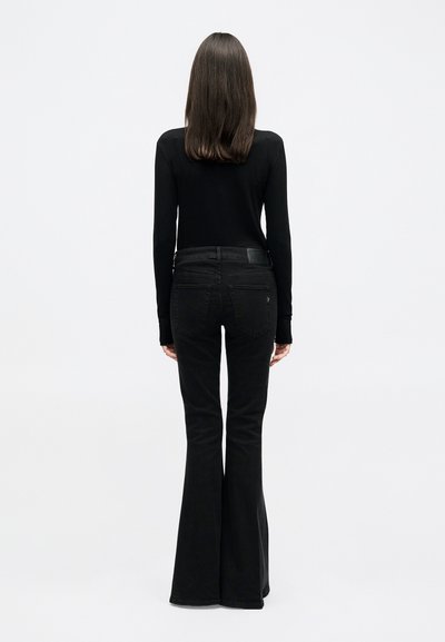 Kobieta stojąca tyłem, ubrana w czarną bluzkę z długim rękawem i czarne rozszerzane dżinsy na jednolitym białym tle.