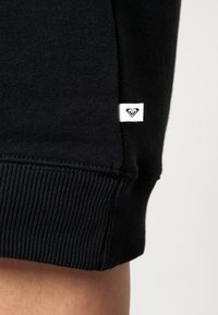 Sweatshirt en coton noir avec poignets côtelés, doté d'une petite étiquette blanche avec un logo cœur. Texture douce et design minimaliste.