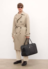 Gabardina beige con botones de doble fila, suéter beige, pantalones crema, bolso de cuero negro y botas marrones. Las gafas añaden un detalle de contraste.