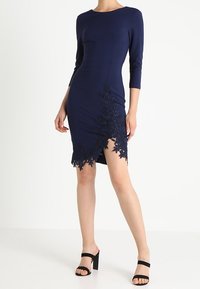 Abito aderente blu navy con maniche a tre quarti, caratterizzato da dettagli in pizzo intricati sull'orlo inferiore e una spaccatura laterale.