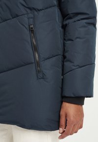 Mørkeblå puffjakke med diagonalt quiltet design, en glidelåslomme på venstre arm og en svart ribbet mansjett på ermet.