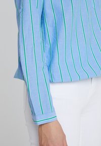 Chemise à manches longues bleu clair avec des rayures verticales vertes, dotée d'un poignet à bouton et d'un tissu doux. Fond blanc visible.