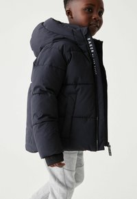 Doudoune marine à capuche, présentant une fermeture éclair bien visible et des accents de logo. Le matériau semble lisse et isolé, renforçant la chaleur.