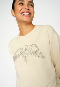 Felpa beige con un design grafico testurizzato in strass che presenta ali e un simbolo. Silhouette morbida e rilassata con scollo rotondo.