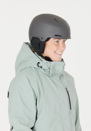 Vrouw in een lichtblauwe winterjas en een grijs sk helmet, die lacht en naar rechts kijkt tegen een witte achtergrond.
