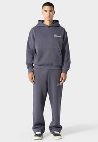 Hedvábná šedá mikina s kapucí z bavlněného směsového materiálu, s přední kapsou a logem na hrudi, spárovaná s odpovídajícími tepláky s logem na stehně.