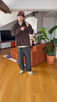 Hnědý fleecový pulovr, světle šedé tričko pod ním, tmavě šedé volné kalhoty a stříbrné boty, stojící v obývacím pokoji s rostlinami.