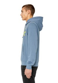Felpa leggera blu con tasca frontale, coulisse e lettere gialle. Tessuto morbido e polsini a coste. Il modello indossa pantaloni neri.