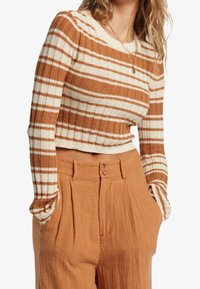 Prążkowany, krótki sweter w brązowo-kremowym kolorze, zestawiony z spodniami z wysokim stanem w rdzawym odcieniu, plisowanymi i o gładkiej fakturze.