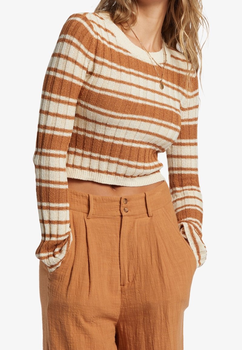 Prążkowany, krótki sweter w brązowo-kremowym kolorze, zestawiony z spodniami z wysokim stanem w rdzawym odcieniu, plisowanymi i o gładkiej fakturze.