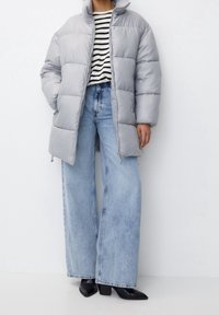 Chaqueta de plumas gris claro con un cuello alto, combinada con una camiseta a rayas en blanco y negro, y jeans de mezclilla azul de pierna ancha, finalizando con botas de tacón negras.